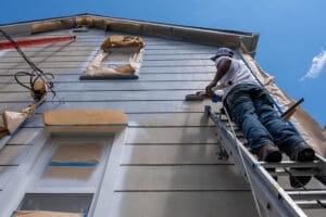 Professional painter on a ladder painting the exterior of a two-story home, illustrating what affects the cost of exterior painting.