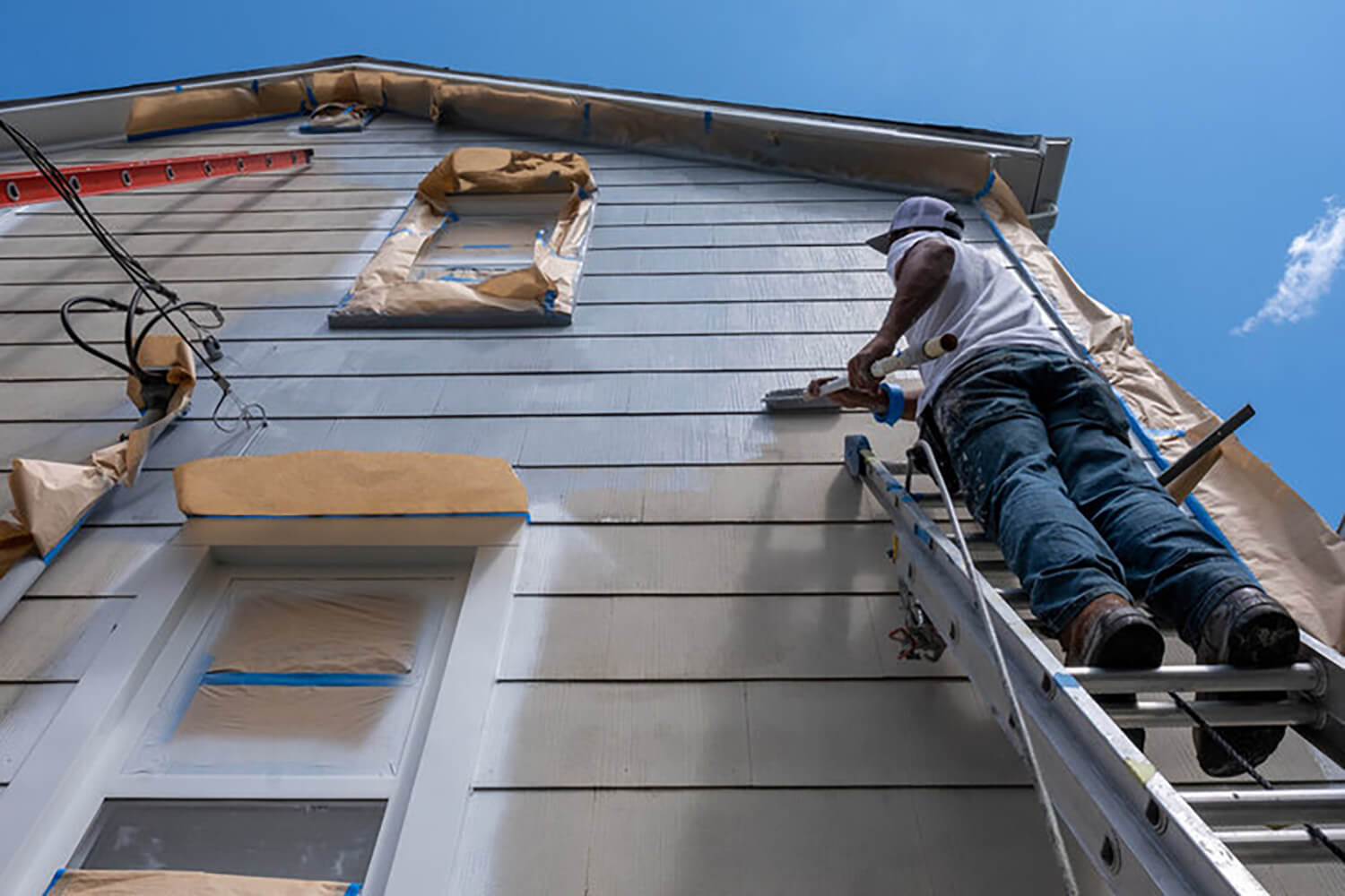 Professional painter on a ladder painting the exterior of a two-story home, illustrating what affects the cost of exterior painting.