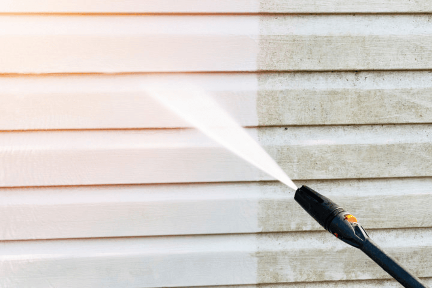 A pressure washer nozzle cleaning dirty siding, showing the contrast between unwashed and freshly cleaned exterior wall surfaces.